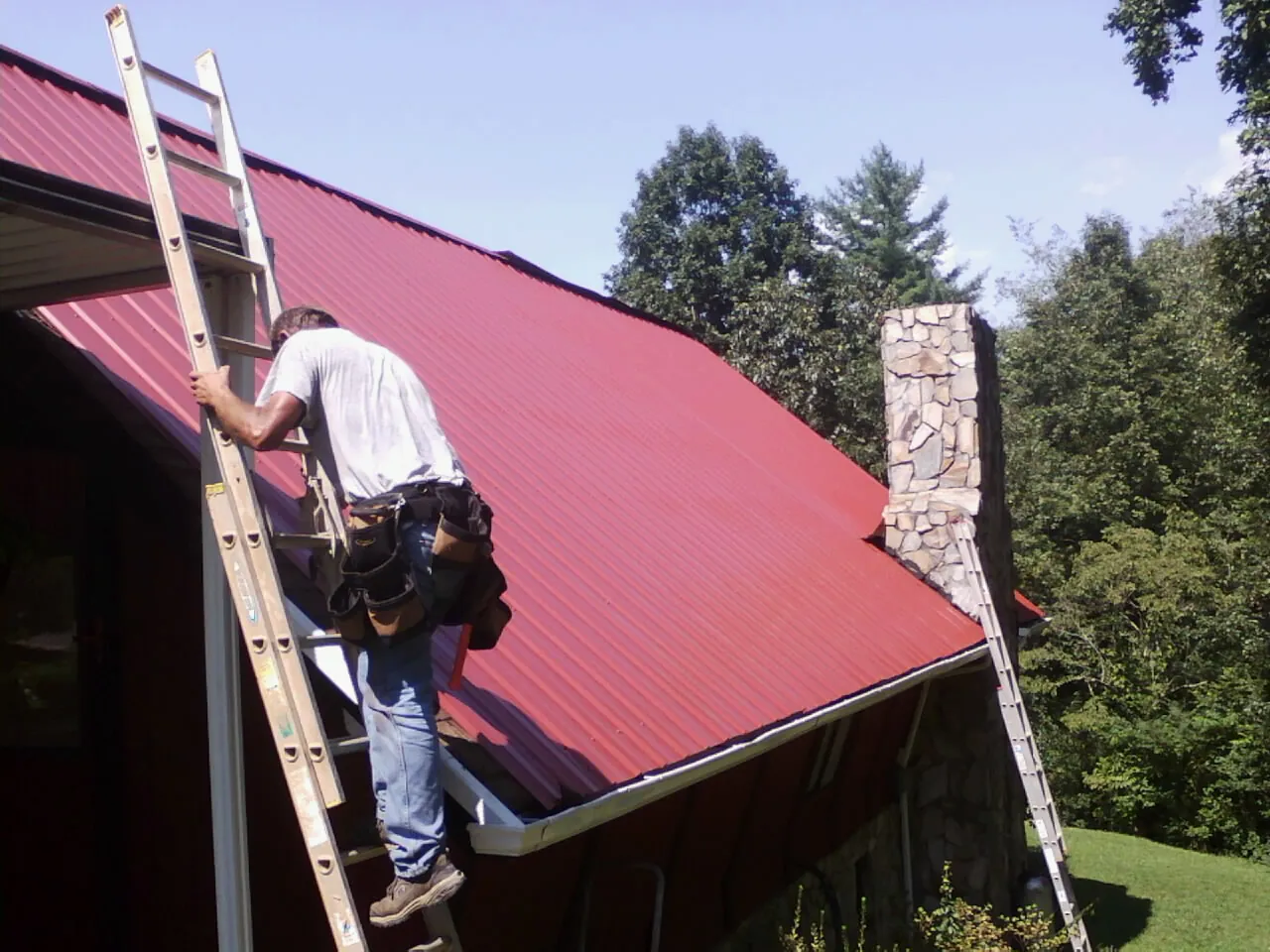 Roofer on a ladder beside a red metal roof and stone chimney — Soffit & Fascia Repair by Roofing Brothers in Babylon